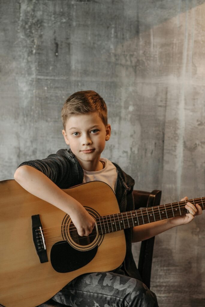 A young boy sits indoors playing an acoustic guitar with a smile.